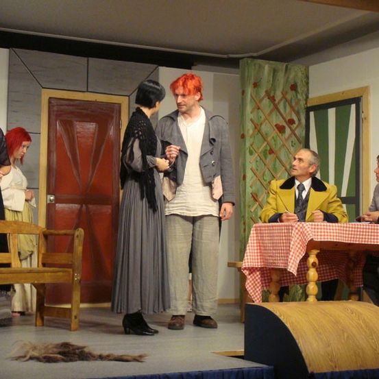 Eine Bühnenaufführung mit Schauspielern in Vintage-Kostümen. Ein Mann und eine Frau in der Mitte interagieren. Eine weitere Frau steht hinter ihnen, und zwei Männer sitzen an einem Tisch.