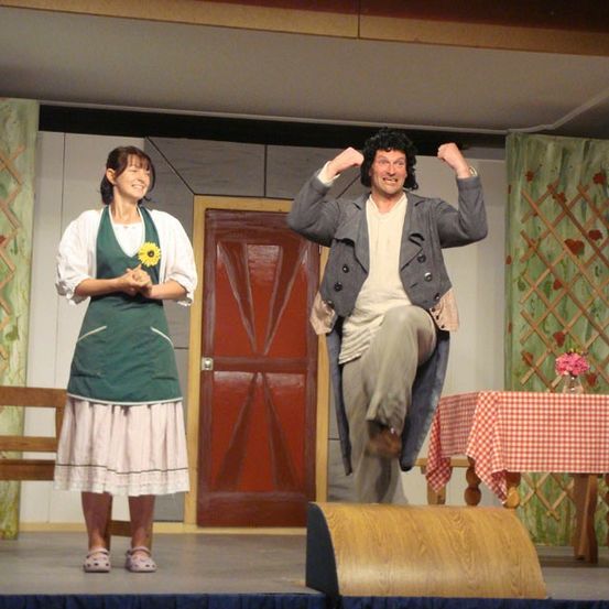 Zwei Darsteller auf der Bühne; eine Frau in Grün steht links und lächelt, während ein Mann in Grau rechts springt. Dahinter eine rote Tür, eine Bank und ein Tisch mit einer rosa Blume.
