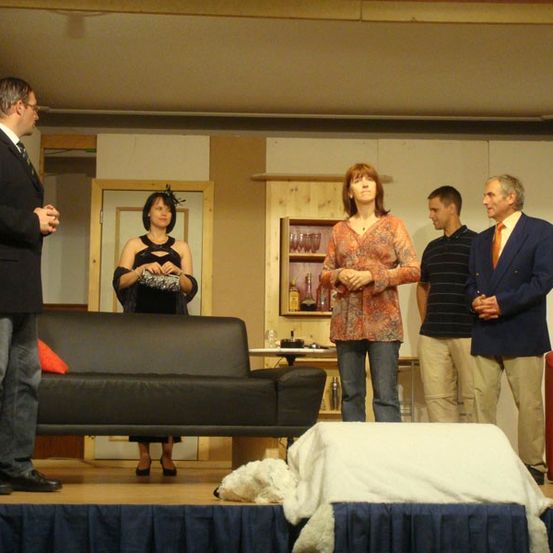 Fünf Personen stehen auf einer Bühne mit einem schwarzen Sofa, einem roten Stuhl und einem Tisch. Eine Frau in der Mitte hält ein silbernes Objekt. Dahinter steht ein Holzschrank mit Weingläsern und Flaschen.