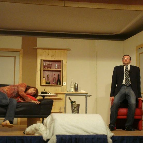 Eine inszenierte Szene zeigt eine Frau, die auf einer Couch liegt, während ein Mann in Anzug und Krawatte in einem roten Stuhl sitzt und sie ansieht. Hinter ihnen steht ein Holzregal mit Flaschen und Gläsern.