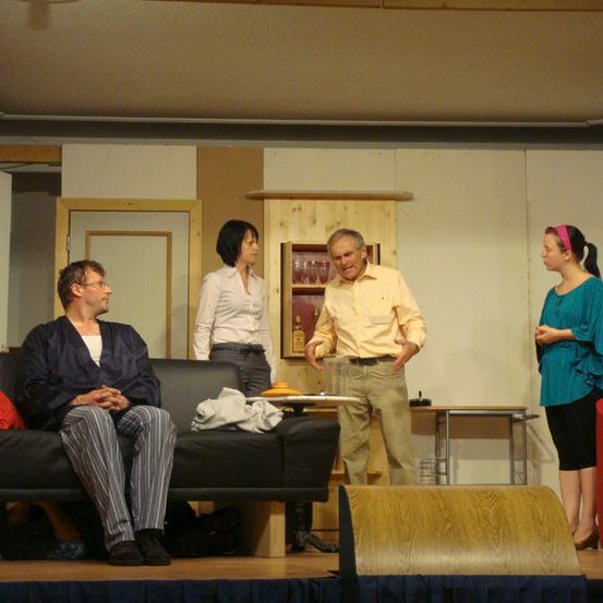 Vier Personen befinden sich auf einer Bühne. Ein Mann sitzt auf einer Couch, ein anderer Mann steht mit den Händen in den Hüften, eine Frau in einem weißen Hemd steht hinter einem Holzschrank und eine Frau in einem blauen Hemd steht rechts.