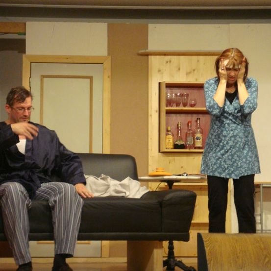 Ein Mann in Pyjama sitzt auf einem Sofa, während eine Frau in einem blauen Kleid neben ihm steht und ihren Kopf hält. Hinter ihnen steht ein Holzregal mit Flaschen und Gläsern, und ein rotes Kissen liegt auf dem Sofa.