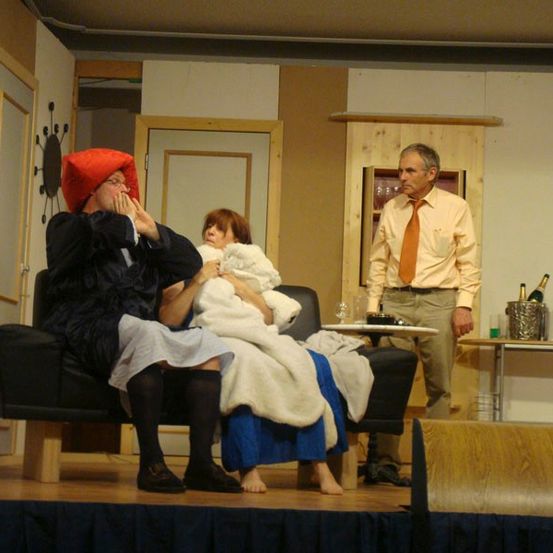 Ein Mann mit einem roten Hut sitzt auf einem Sofa mit einer Frau. Ein weiterer Mann steht hinter ihnen in der Nähe eines Tisches mit Flaschen.