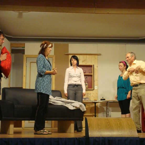 Fünf Personen stehen auf einer Bühne, möglicherweise bei einer Aufführung. Sie stehen im Halbkreis, mit einer Couch und einem Tisch im Hintergrund.