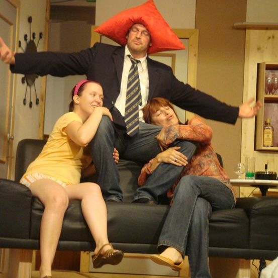 Ein Mann im Anzug mit einem roten Kissen auf dem Kopf sitzt auf einem Sofa mit zwei Frauen, eine in einem gelben Shirt und die andere in einer Bluse mit Blumenmuster.
