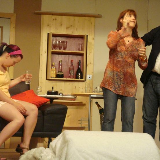 Drei Personen sind auf einer Bühne; eine Frau in Gelb hält ein Glas, eine Frau in Orange lacht und ein Mann im Anzug lehnt sich an sie. Dahinter steht ein hölzerner Schrank mit Spirituosenflaschen und Gläsern.