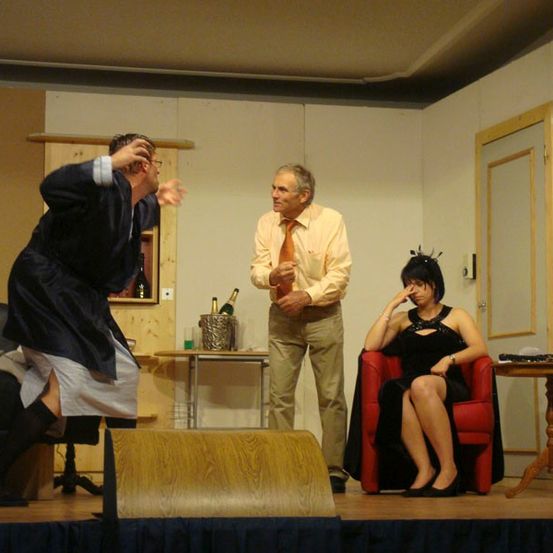 Drei Personen auf einer Bühne. Ein Mann steht mit verschränkten Armen, ein anderer sitzt in einem Stuhl und eine Frau steht links. Hinter dem stehenden Mann befindet sich ein Holztisch mit einer Flasche und einem Eimer.