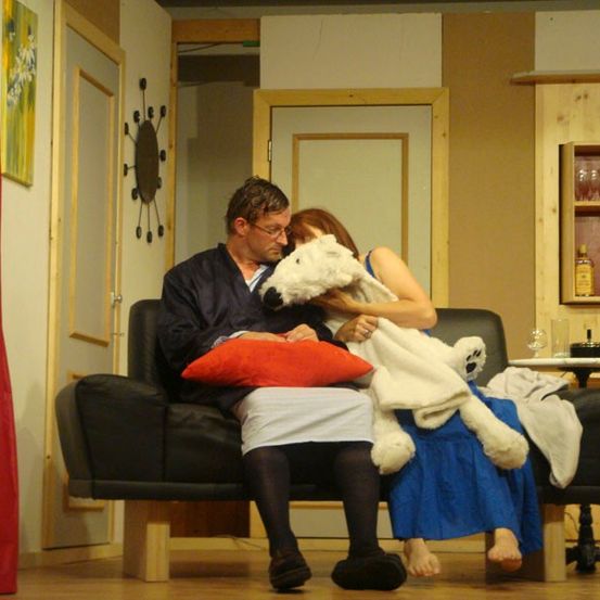 Ein erwachsener Mann und eine Frau sitzen mit einem großen weißen Hund auf einer Couch und halten eine Decke. Sie sind in einem Raum mit einem roten Vorhang, einer Uhr und einem Holzschrank mit Flaschen.