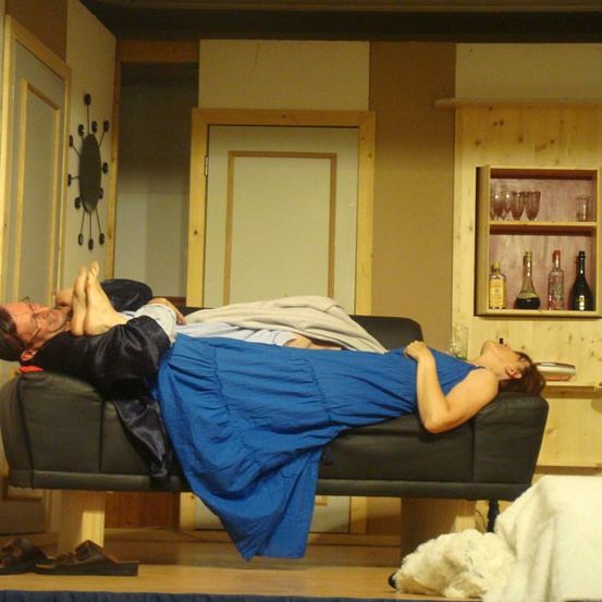 Zwei Personen liegen auf einer Couch in einer inszenierten Umgebung mit einem Holzschrank und Regalen im Hintergrund.