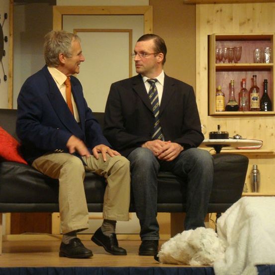 Zwei Männer in Anzügen sitzen auf einer Couch. Einer trägt eine Krawatte und hat die Hände verschränkt, während der andere die Hände auf den Knien hat. Sie lächeln beide.