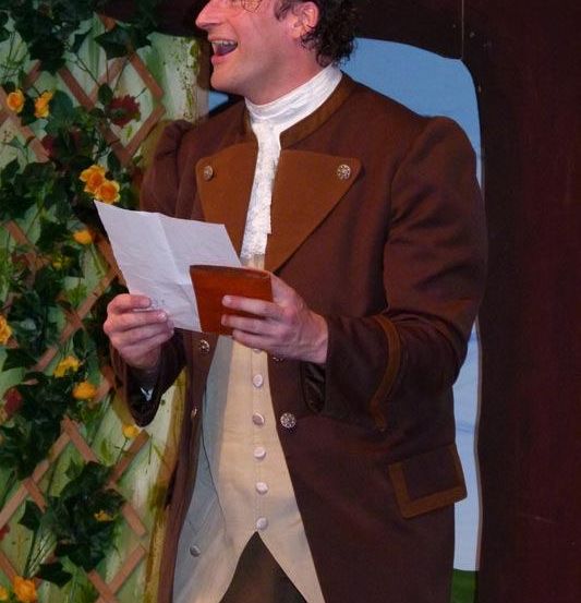 Ein Mann in einem braunen Mantel und weißer Weste hält ein Stück Papier und eine Geldbörse, lächelt auf der Bühne.