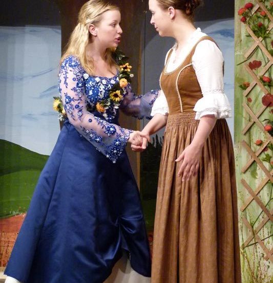 Zwei Frauen in historischen Kostümen halten auf einer Bühne Händchen. Die Frau auf der linken Seite trägt ein blaues Kleid mit Sonnenblumen. Der Hintergrund zeigt eine bemalte Szene mit grünen Bäumen und roten Blumen.