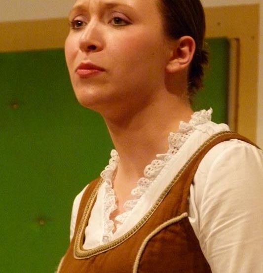 Eine Frau in einem braun-weißen Kleid steht und schaut mit ernster Miene zur Seite.