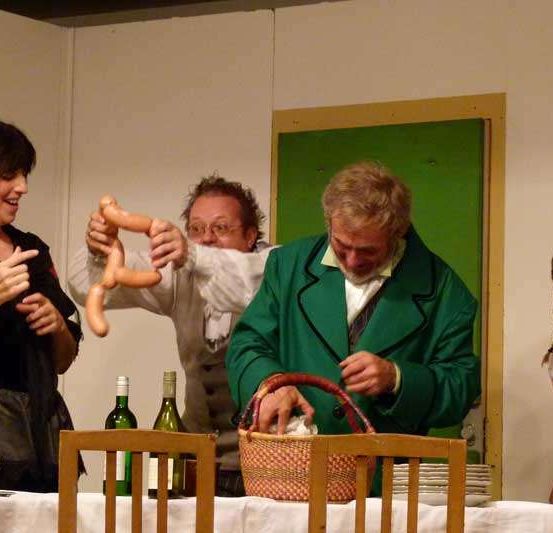 Eine Gruppe von Menschen versammelt sich um einen Tisch mit Flaschen und einem Korb, ein Mann hält eine Wurst.