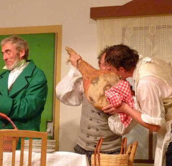 Drei Männer in historischer Kleidung, einer hält einen Korb und die anderen bereiten sich darauf vor, ein großes Stück Käse zu probieren.