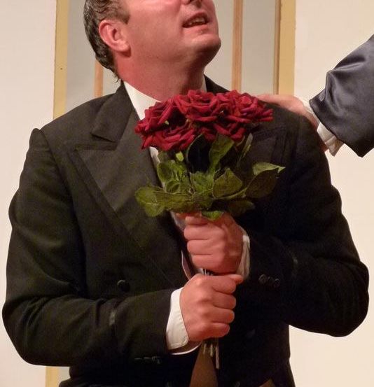 Ein Mann in einem schwarzen Anzug hält einen Strauß roter Rosen und schaut mit offenem Mund nach oben.