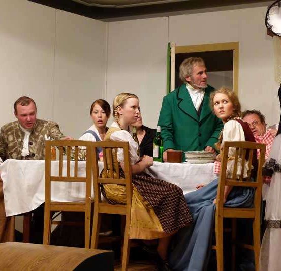 Eine Gruppe von Menschen in historischer Kleidung sitzt um einen Tisch herum, wobei eine Person steht und mit ihnen spricht.