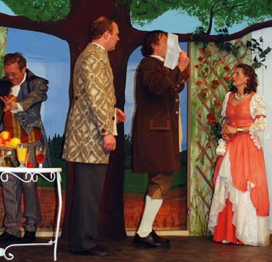 Fünf Personen stehen auf einer Bühne, einer Mann hält ein weißes Tuch. Zwei Frauen und zwei Männer stehen. Eine Frau hält einen Fächer. Ein Tisch mit Getränken ist hinter ihnen.