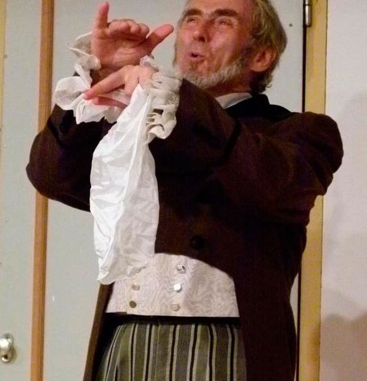 Ein älterer Mann in historischer Kleidung mit einem gerüschten Hemd und gestreiften Hosen gestikuliert dramatisch mit einem Stück Papier in den Händen.