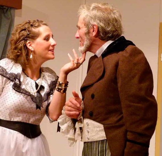 Ein älterer Mann in einem braunen Mantel unterhält sich mit einer Frau in einem weißen Kleid und einem schwarzen Gürtel, beide sehen ernst aus.