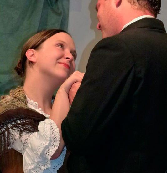 Eine Frau in einem historischen Kleid lächelt, während ein Mann in einem Anzug ihre Schulter hält.