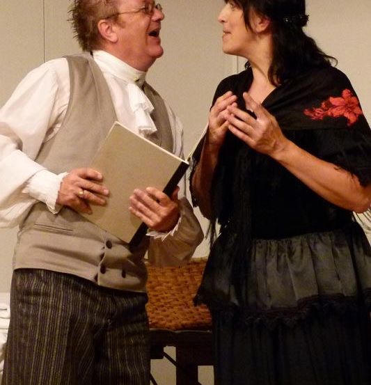 Ein Mann in einem weißen Hemd und Weste hält ein Buch und spricht zu einer Frau in einem schwarzen Kleid mit einer roten Blume.