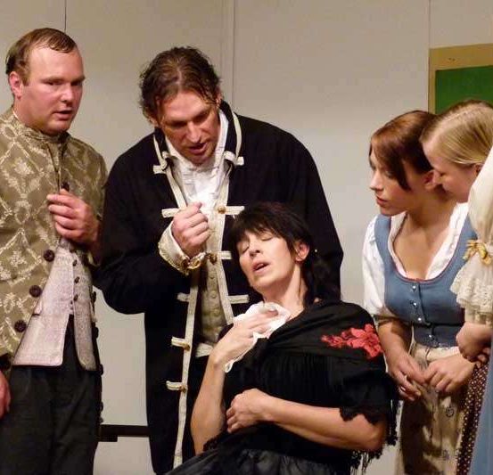 Fünf Personen stehen auf der Bühne, zwei Männer und drei Frauen. Die Frau in der Mitte sitzt und hält ein weißes Tuch. Der Mann rechts von ihr schaut sie an. Der Mann links beobachtet sie.