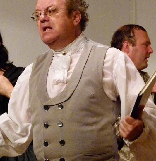 Ein Mann mit lockigem Haar und Brille singt und hält ein Buch in der Hand, er trägt ein weißes Hemd und eine graue Weste.