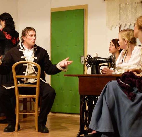 Ein Mann in historischer Kleidung sitzt und gestikuliert, während er mit einer Gruppe von Frauen spricht, die um einen Tisch mit einer Nähmaschine sitzen.