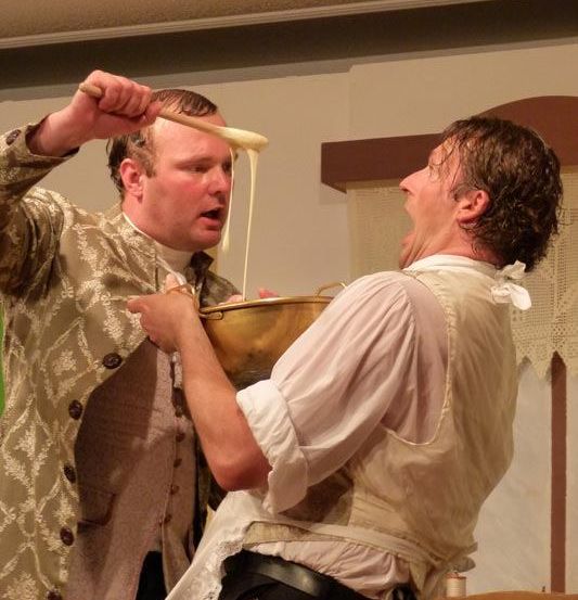 Zwei Männer führen eine Szene auf; einer gießt Sauce in eine Schüssel, die vom anderen gehalten wird, der lacht. Sie tragen historische Kleidung.