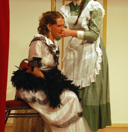 Zwei Frauen in Vintage-Kleidung, eine sitzt auf einem Stuhl, die andere steht hinter ihr. Die stehende Frau richtet die Haare der sitzenden Frau.