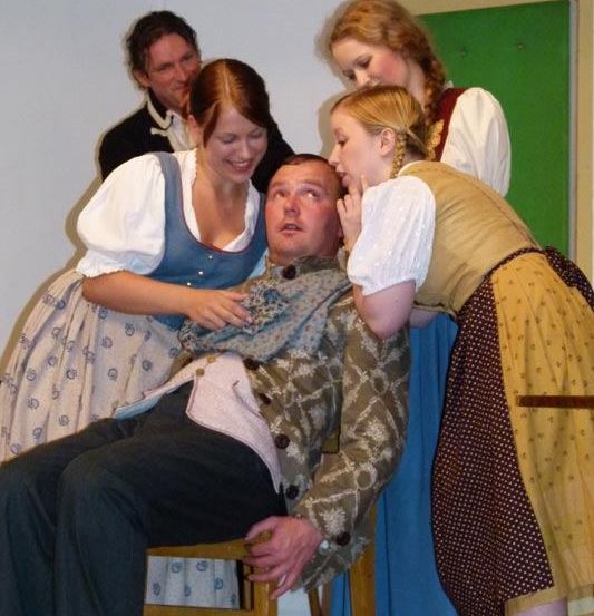 Ein Mann auf einem Stuhl, umgeben von Frauen in traditioneller Kleidung, möglicherweise bei einer Theateraufführung, mit einem grünen Hintergrund hinter ihnen.