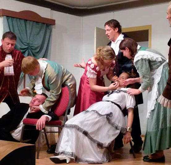 Eine Gruppe von Menschen ist auf der Bühne, einige liegen auf dem Boden und andere stehen. Der Mann in der Mitte liegt auf einem Stuhl. Eine der Frauen beugt sich vor und scheint den Mann zu überprüfen. Die Menschen sind in Kostümen gekleidet.