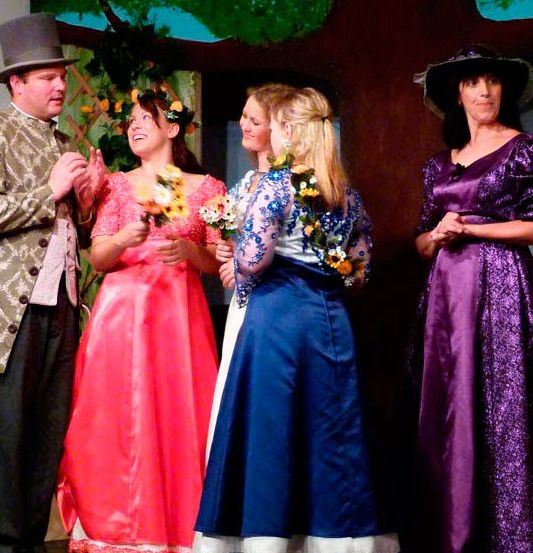 Fünf Menschen in Kostümen stehen auf einer Bühne, wahrscheinlich für ein Theaterstück. Eine Frau hält Blumen.