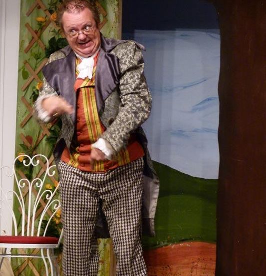 Ein Mann in einem bunten Kostüm steht lächelnd auf der Bühne. Er steht neben einem weißen Stuhl und einem Baum, mit einer landschaftlichen Kulisse im Hintergrund.