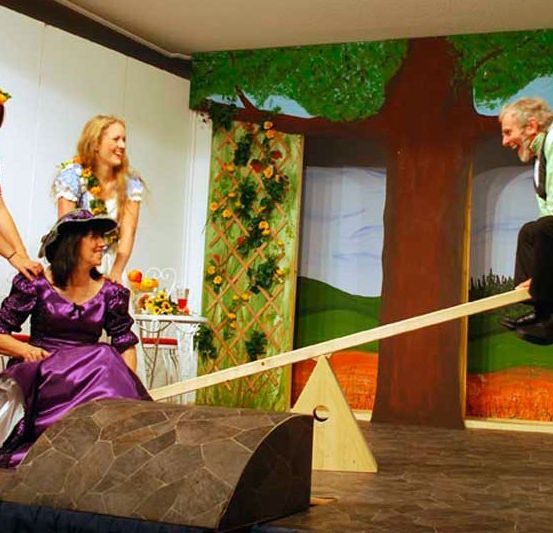 Eine Gruppe von Schauspielern führt eine Szene auf, bei der einer auf einer Wippe sitzt, eine Frau die Basis der Wippe hält und eine weitere Frau in einem lila Kleid lächelt.