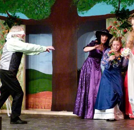 Auf der Bühne deutet ein Mann mit einem Blindfold, während drei Frauen, eine in einem lila Kleid, eine andere in Blau und die dritte in Weiß, um einen Baumkulisse stehen.