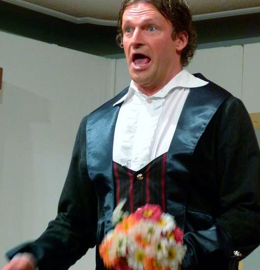 Ein Mann in formeller Kleidung mit einem Blumenstrauß tritt auf der Bühne auf.