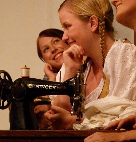 Drei Frauen sitzen neben einer Singer-Nähmaschine, lächeln und plaudern. Die Frau in der Mitte hat geflochtene Haare.