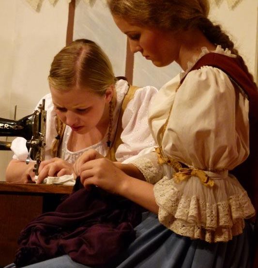 Zwei Frauen in Vintage-Kleidung arbeiten an einer Nähmaschine, eine konzentriert sich auf das Nähen, die andere beobachtet.