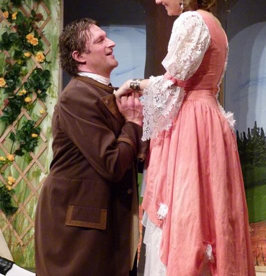 Ein Mann in einem braunen Mantel kniet und hält die Hand einer Frau, beide lächeln. Die Frau trägt ein rosa Kleid mit Spitze. Sie stehen auf einer Bühne mit einer Rankhilfe und grünem Laub im Hintergrund.