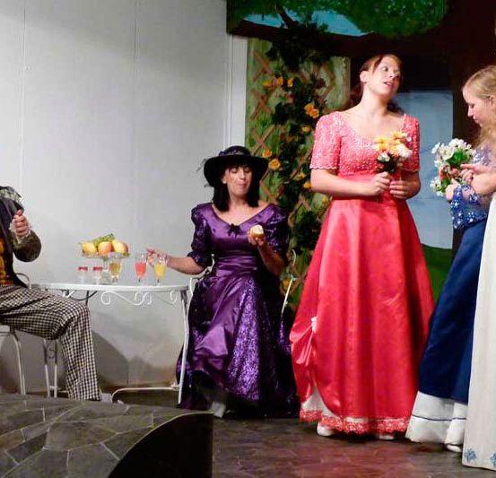 Eine Gruppe von Darstellern auf der Bühne, darunter ein Mann im Anzug, zwei Frauen mit Blumen und eine Frau in einem lila Kleid. Ein Tisch mit Getränken ist im Vordergrund.