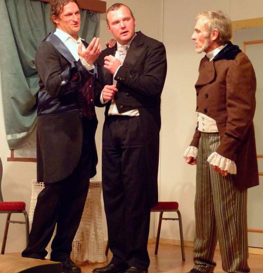 Drei Männer in formeller Kleidung stehen auf einer hölzernen Bühne. Der Mann in der Mitte gestikuliert mit seinen Händen, während die anderen aufmerksam zuhören.