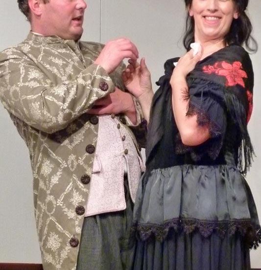 Ein Mann und eine Frau in Kostümen aus der Vergangenheit lächeln und schauen sich an. Der Mann hält ein kleines Objekt und die Frau einen weißen Stoff.