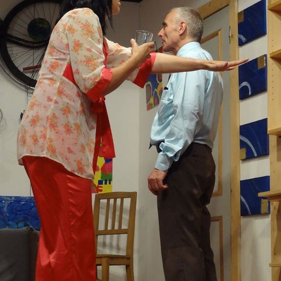 Eine Frau in einem Blumenkleid hält ein Glas und streckt ihren Arm in Richtung eines älteren Mannes in einem hellblauen Hemd und braunen Hosen. Sie stehen in einem Raum mit einem Holzstuhl und farbenfrohen Gemälden an der Wand.