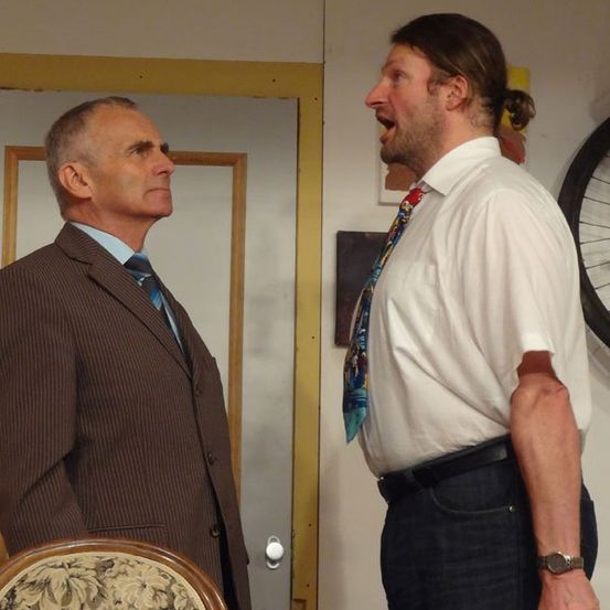 Zwei Männer in formeller Kleidung stehen einander gegenüber, einer trägt einen braunen Anzug, der andere ein weißes Hemd mit einer gemusterten Krawatte. Der Mann in Weiß scheint zu schreien.