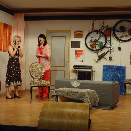 Zwei Frauen in einem Raum, eine telefoniert. Fahrräder hängen an der Wand. Ein abgedecktes Sofa und ein Tisch sind in der Nähe.