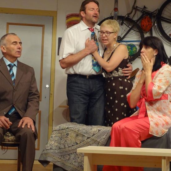 Vier Menschen sind in einem Raum versammelt. Ein Mann sitzt auf einem Stuhl, während eine Frau in einem Blumenkleid auf einem Bett sitzt. Eine weitere Frau mit Brille steht hinter einem Mann mit Krawatte, der zu sprechen scheint.