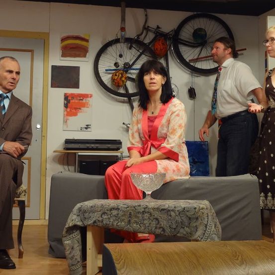 Eine Gruppe von vier Personen in einem Raum. Ein Mann im Anzug sitzt auf einem Stuhl, während eine Frau in einem Kleid auf einer Couch sitzt. Ein weiterer Mann in einer Krawatte steht neben ihr, und eine Frau mit Brille steht neben ihm. Fahrradreifen sind an der Wand montiert.