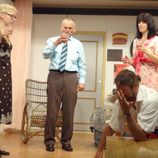 Vier Personen stehen in einem Raum. Zwei Frauen trinken aus Gläsern, ein Mann trinkt und der andere Mann hält sich den Kopf in den Händen. Hinter ihnen steht ein Stuhl und eine Tür.